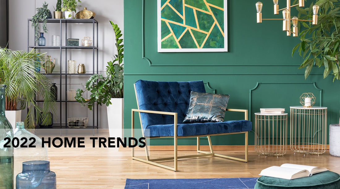 A room with emerald green wall, wired end tables, velvet blue and bronze chair, various plants, and an open metal bookshelf with vases and plants and a gold industrial chandelier.